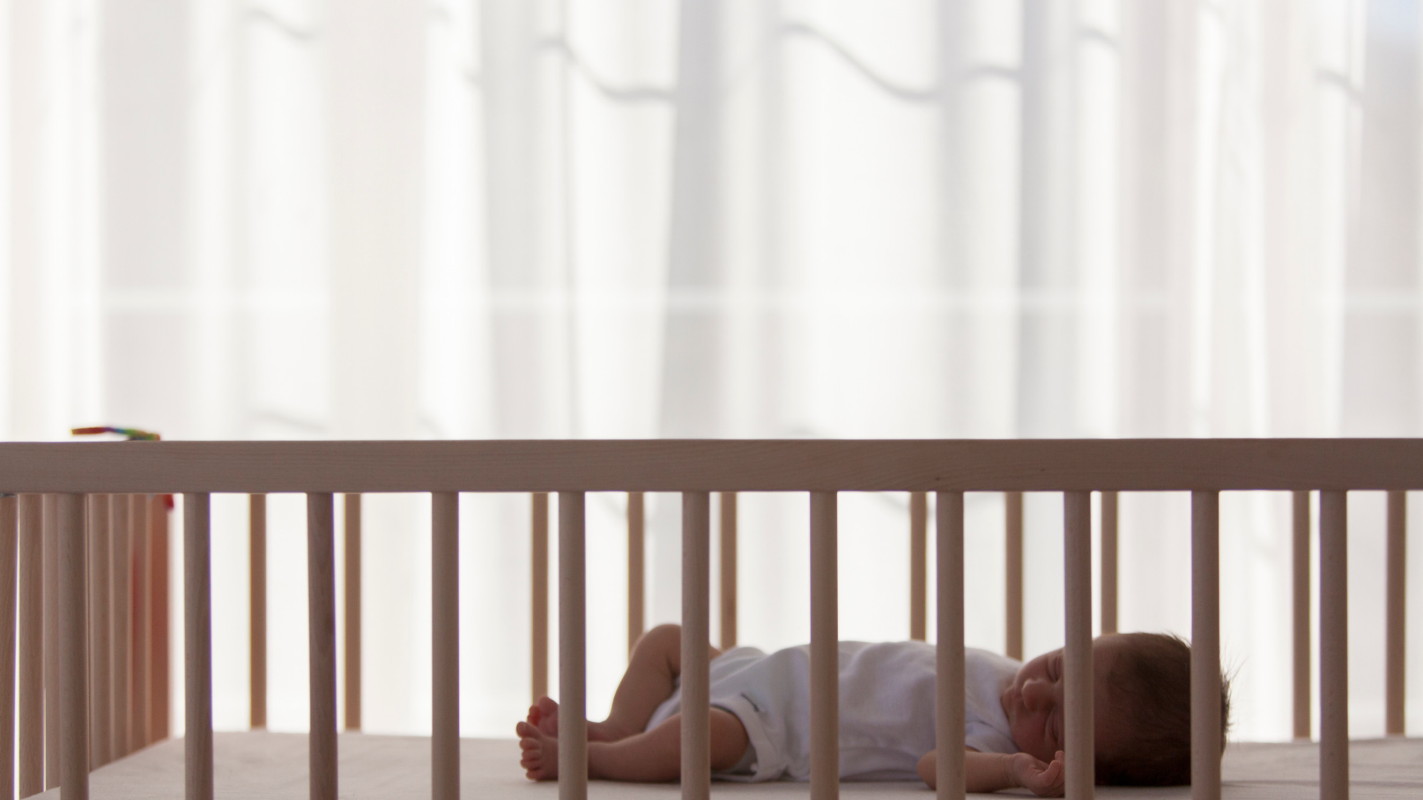 A newborn baby sleeping in a wooden crib in front of a window.